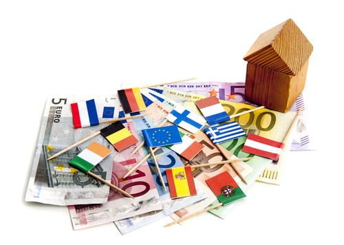 A photo of a little wooden house, and lots of different types of money from all around the world, with different country's flags sitting on top.