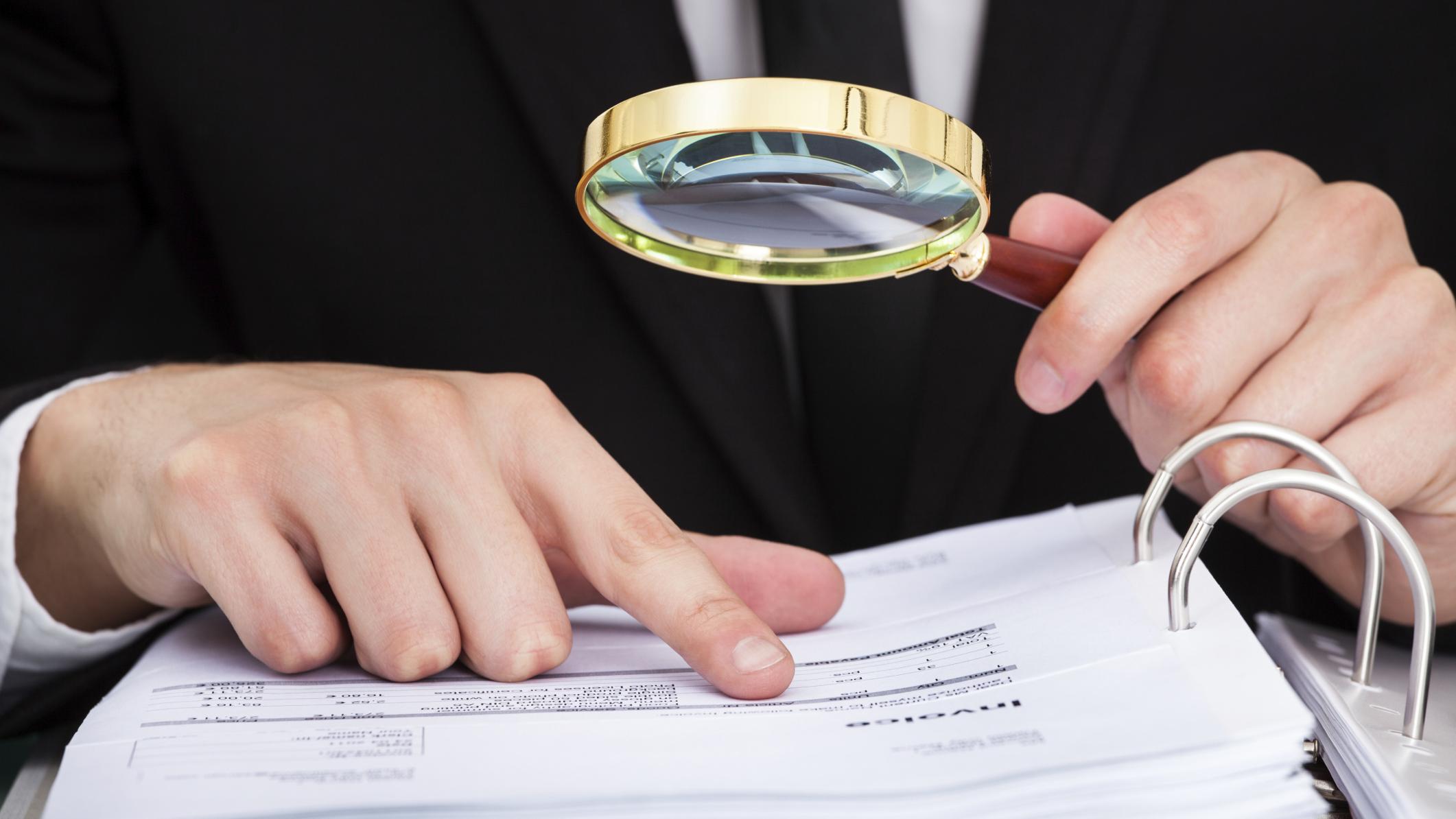 Photo of a man in a business suit using a magnifying glass, marking his place on a document with his right index finger.