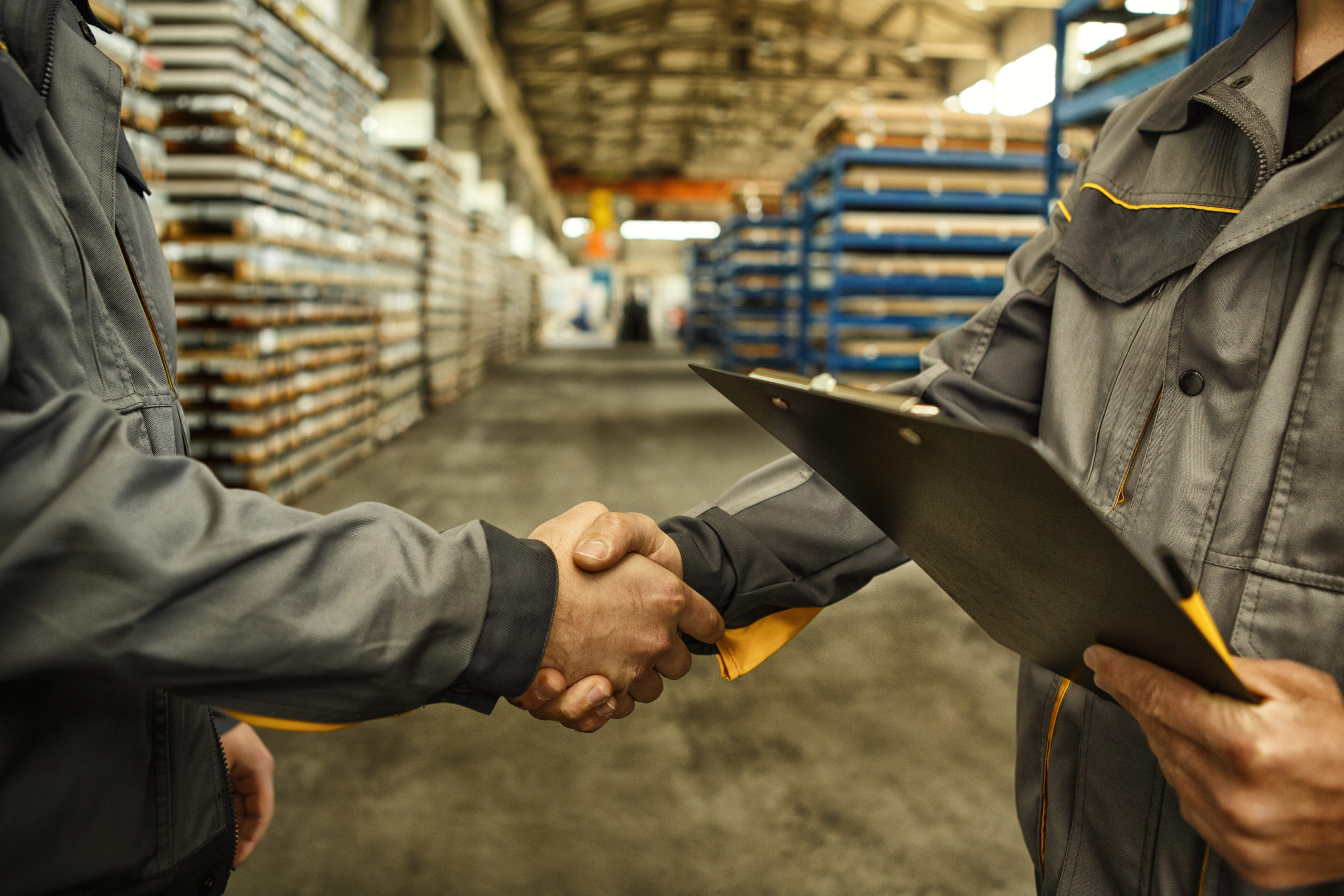 Cropped close up of a worker holding clipboard shaking hands with his colleague at an industrial factory.