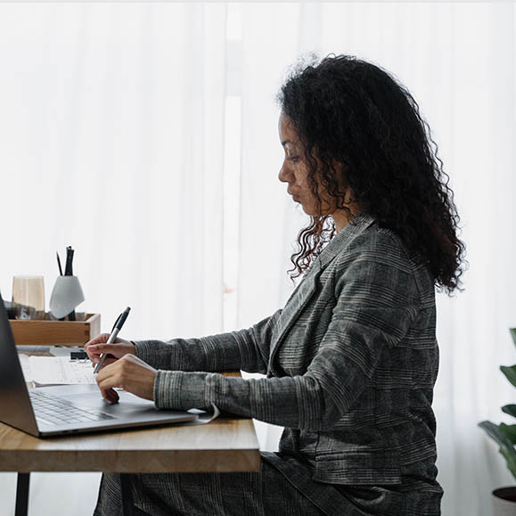 Photo of a woman sitting in front of an open laptop, holding a pen.