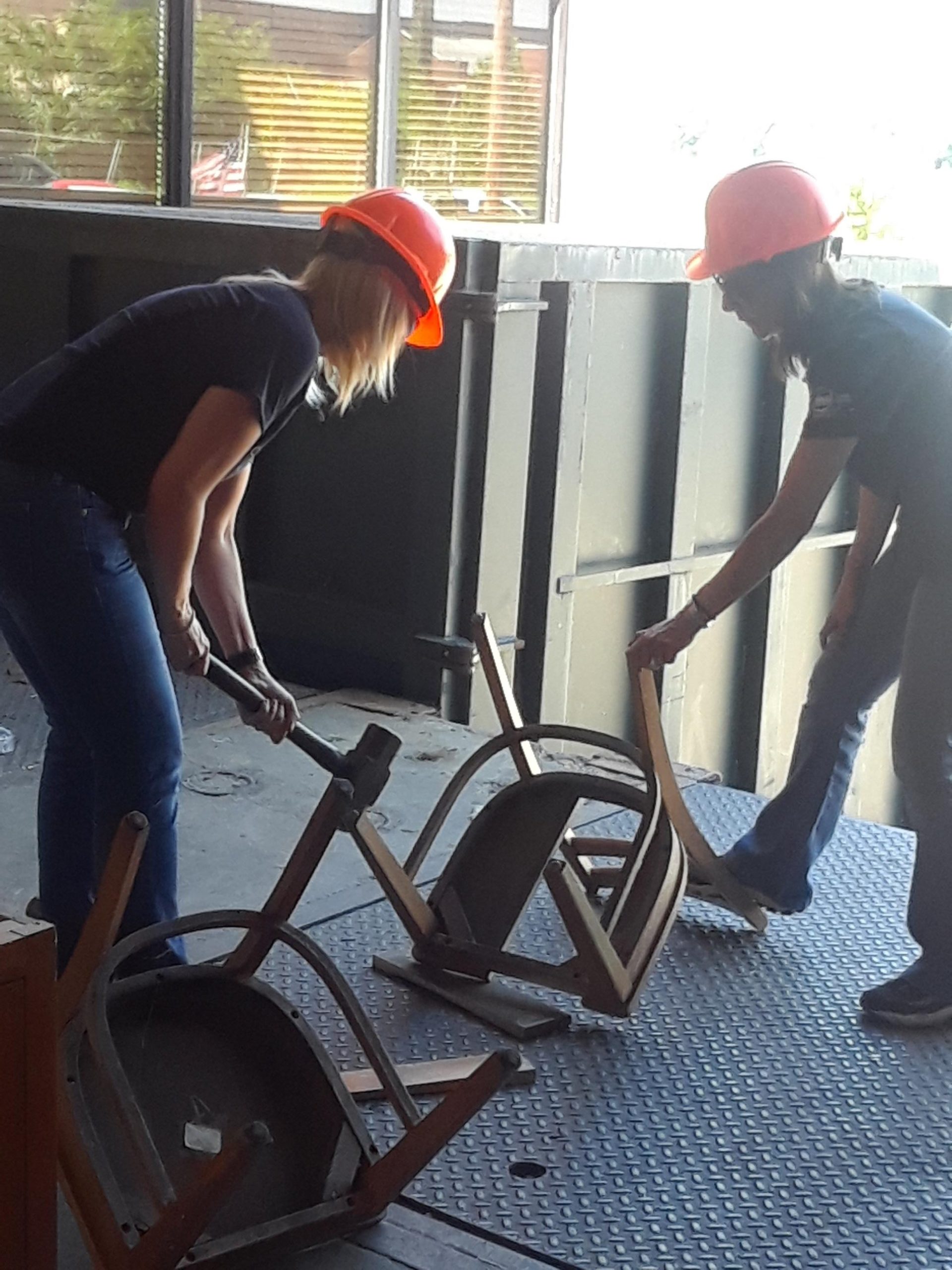 Photo of two people, wearing orange hardhats, taking a sledgehammer to an upside-down, half-destroyed chair.