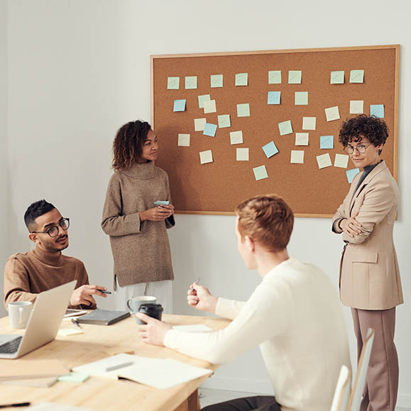 A tan color-schemed photo of a team discussing things, with a board of post-it notes in the background.